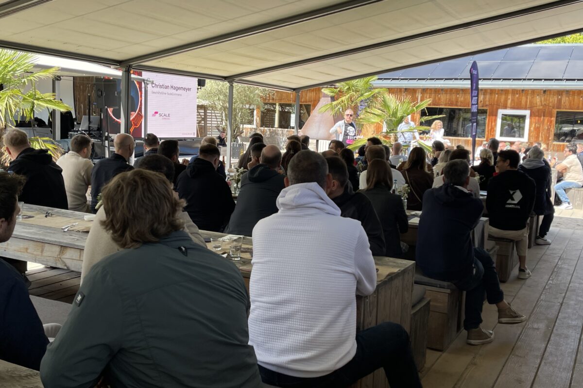 Die Besucher des BBQ sitzen an Holzbänken draußen und hören dem Speaker zu.